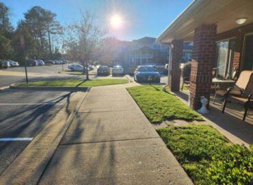 Sidewalk next to grass and parking lot. Sun shines brightly. Brick building with chairs on right.