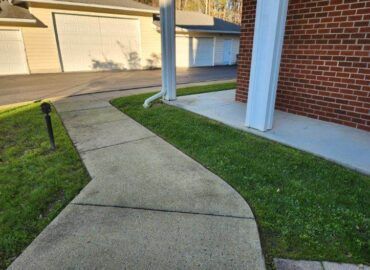 Concrete walkway curving alongside green grass, a red brick building, and garages in the background.