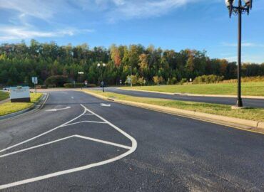 Asphalt road with lane markings leading to a forest. Blue sky, grassy areas, and a lamppost.