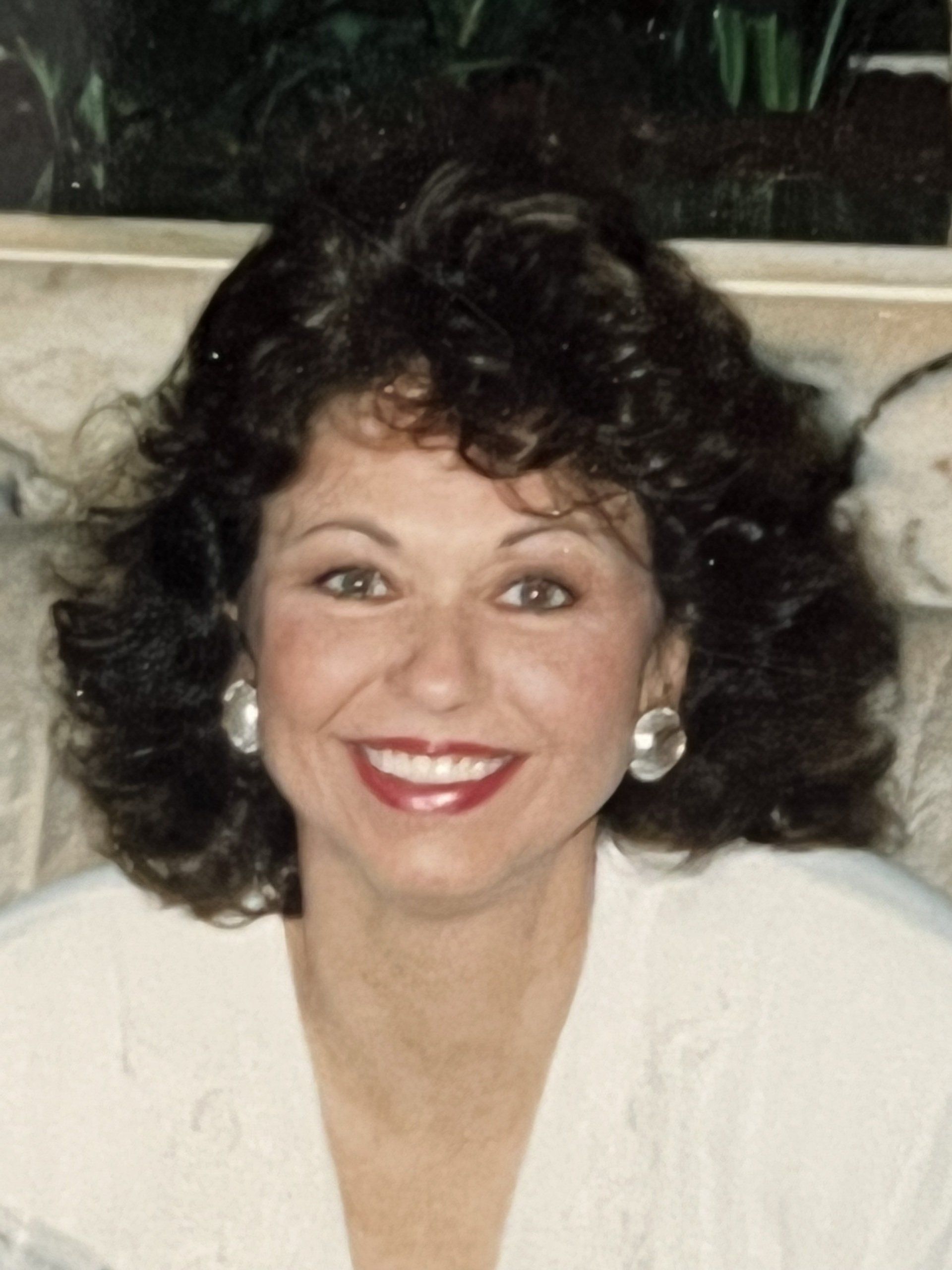 Smiling woman with dark curly hair, red lipstick, and large earrings against a softly lit indoor background