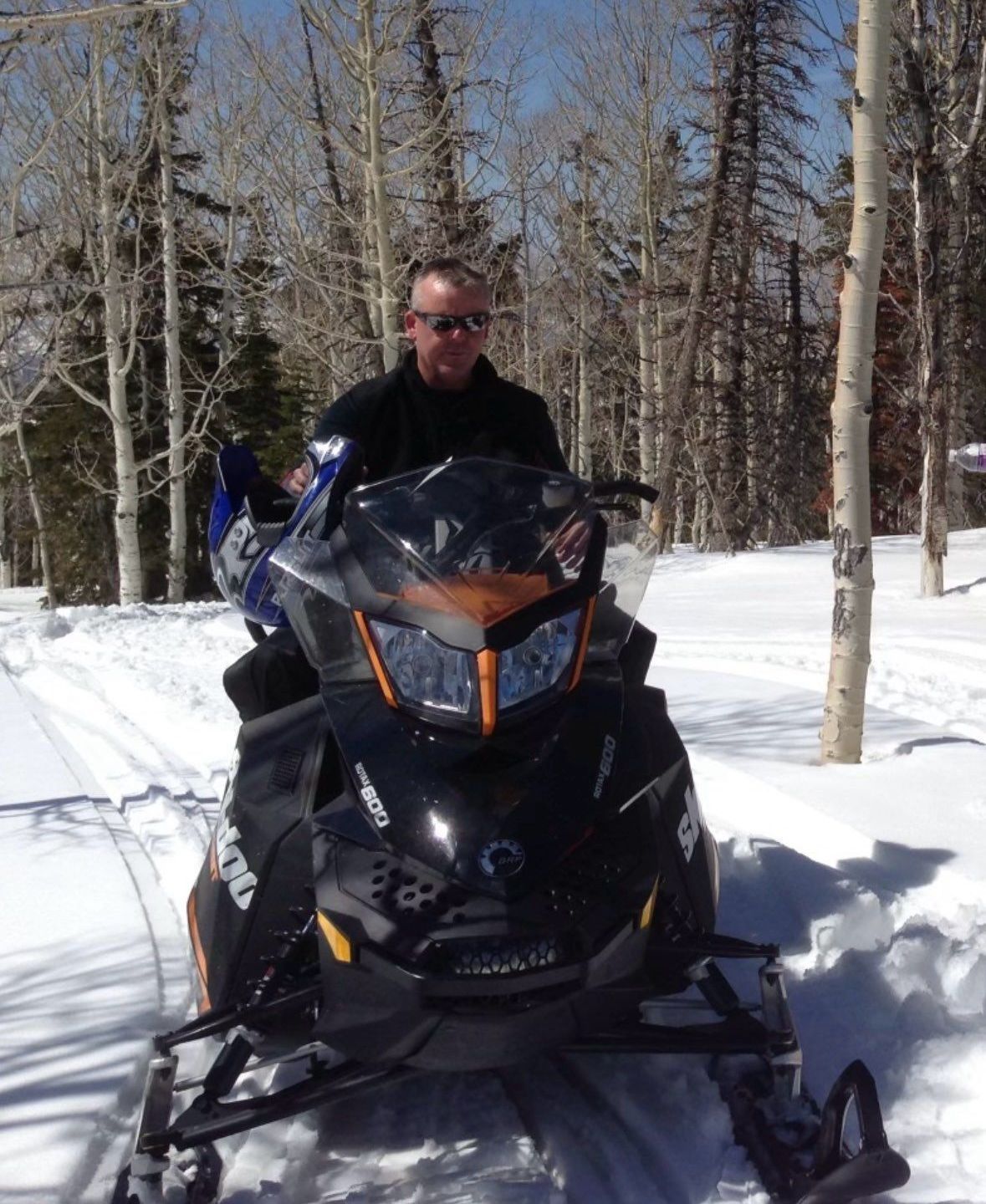 Person on a snowmobile in a snowy forest, wearing dark clothing and sunglasses.