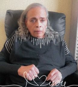 Elderly woman sitting in a black chair, wearing a dark sweater with white fringe, hands resting in her lap.