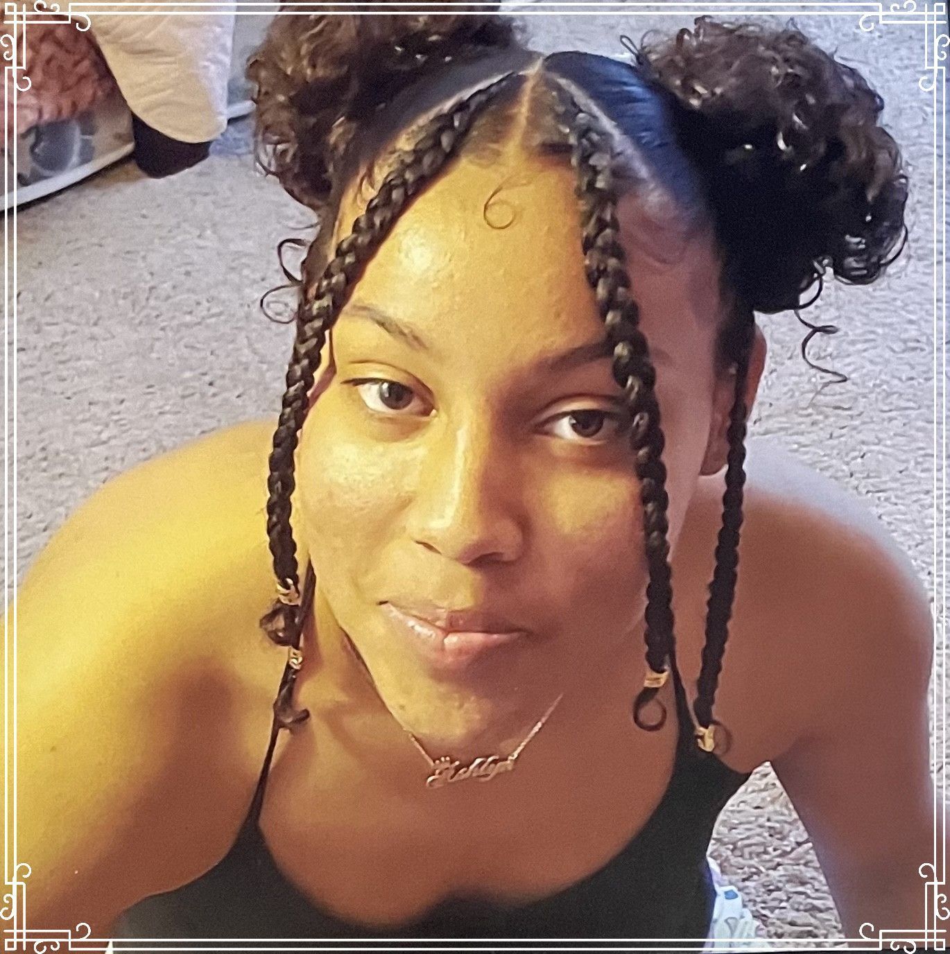 Person with braided hair and curly buns takes a selfie indoors, wearing a black top and necklace.