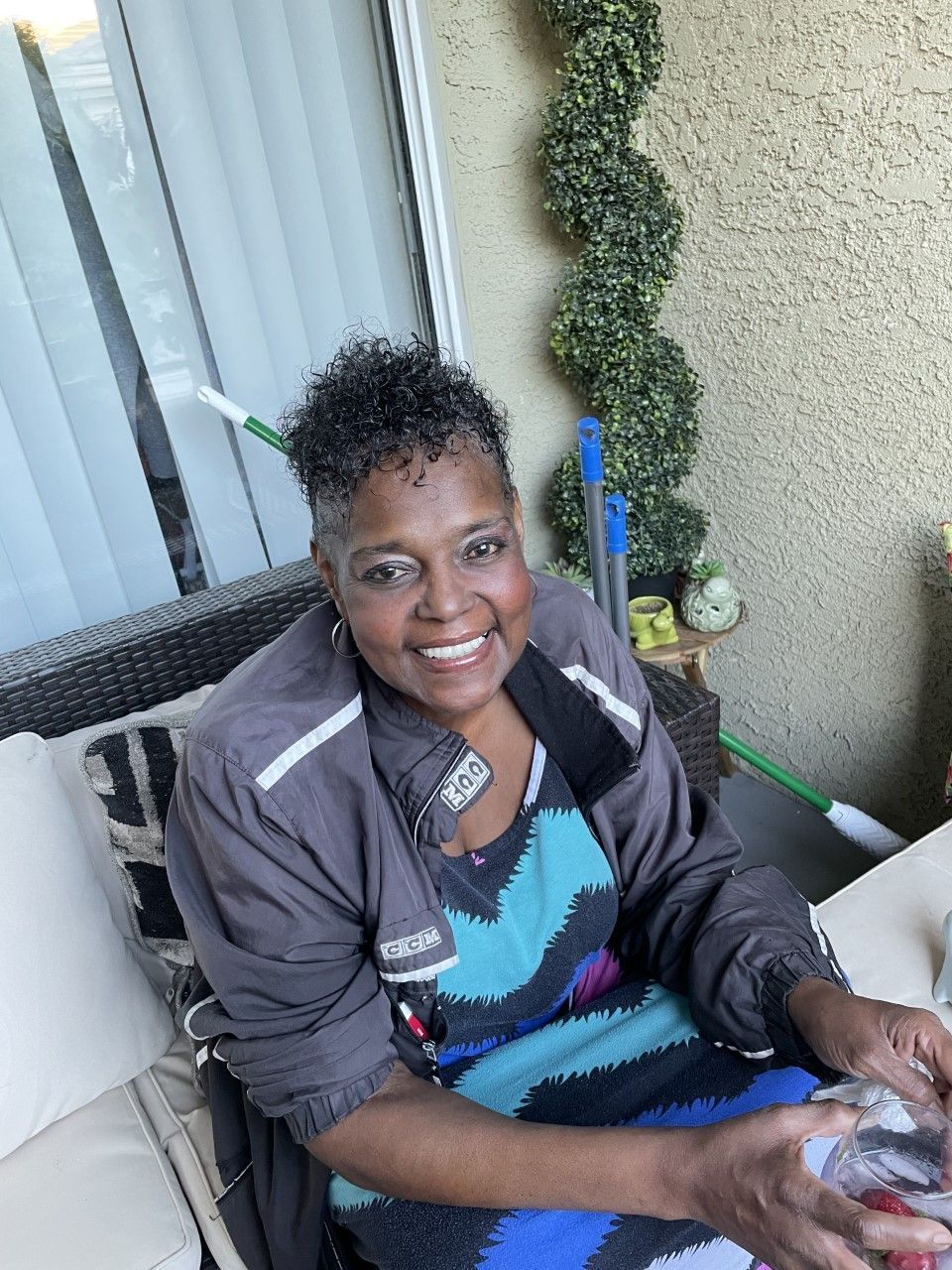 Smiling person seated outdoors on a porch, holding a small pet in their lap, beside a coiled green plant decor