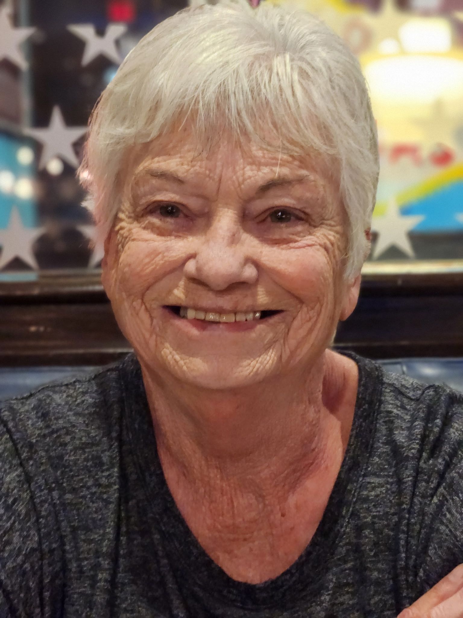 Smiling woman with short white hair in a dark top, seated indoors against a blurred American flag background