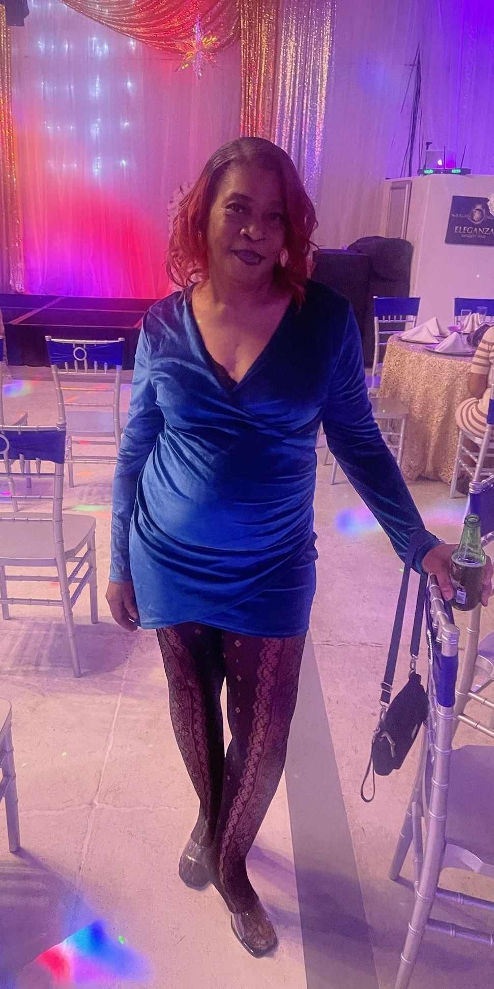 Woman in a blue dress standing at a decorated indoor event with purple lighting, holding a bag.