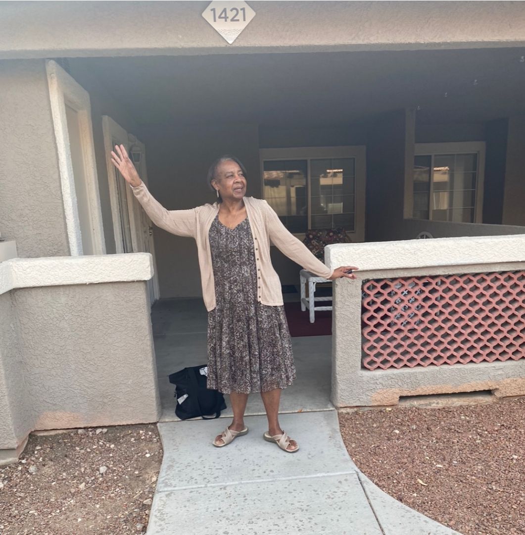 Woman standing at a front porch, waving with one arm raised, beside a house entrance with number 1421.