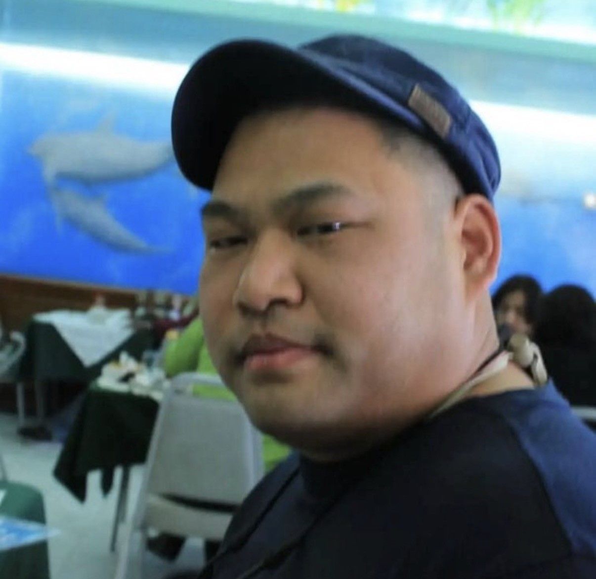 Man in a navy cap and shirt seated in a dining area, looking toward the camera.