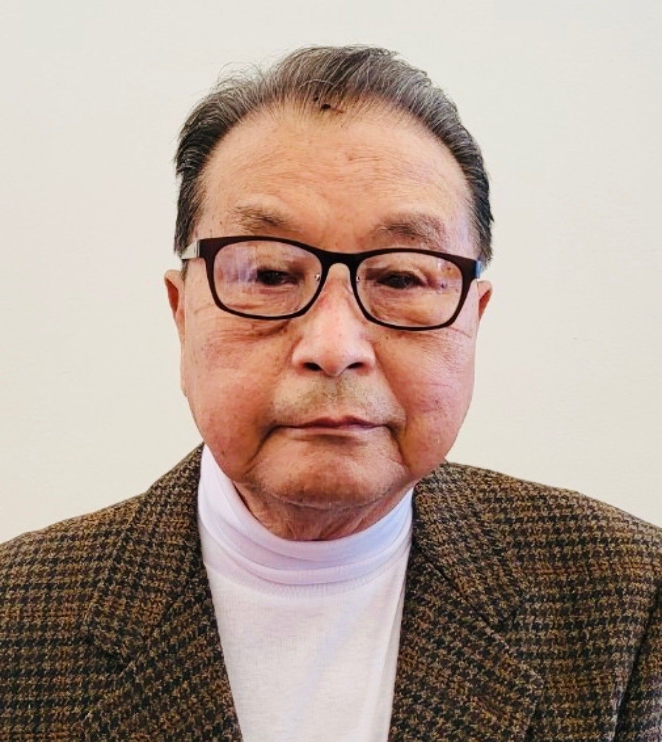 Portrait of a man wearing glasses, a brown plaid jacket, and a white turtleneck against a plain background