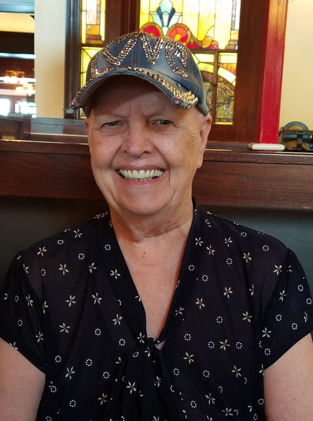 Smiling person in a patterned cap and dark shirt seated indoors with stained glass behind them