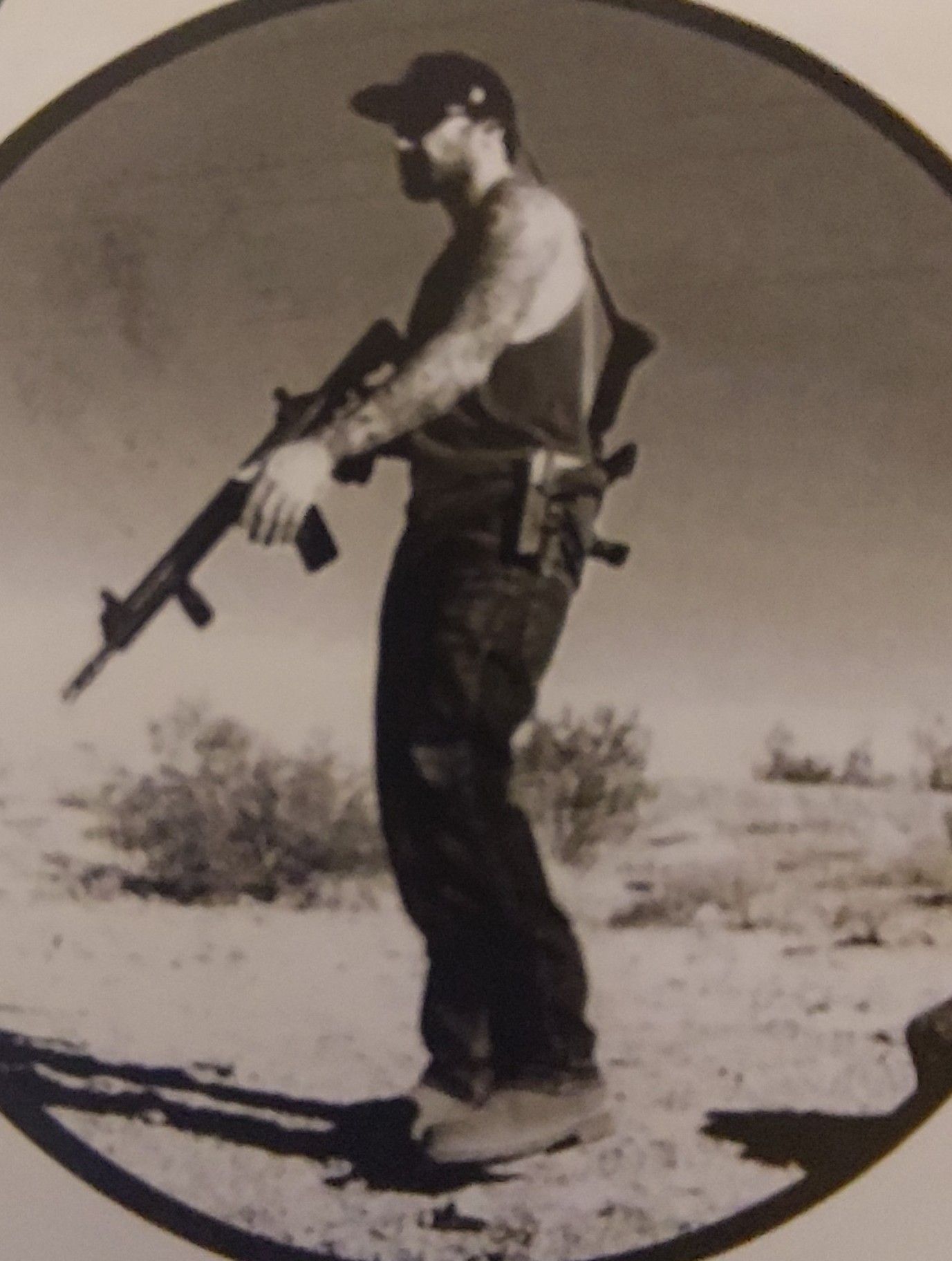 Man holding a rifle in a desert setting, standing in profile under a bright sky.