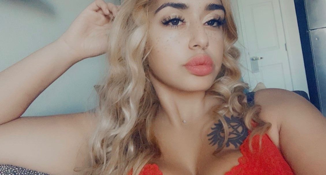 Woman in a red top posing with one hand behind her head, blonde hair, and a floral shoulder tattoo.