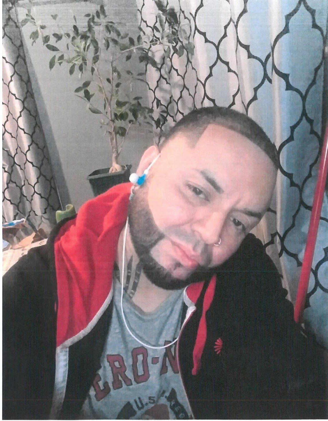 Man in a red hoodie and black jacket, tilting his head with a slight smile indoors near patterned curtains.