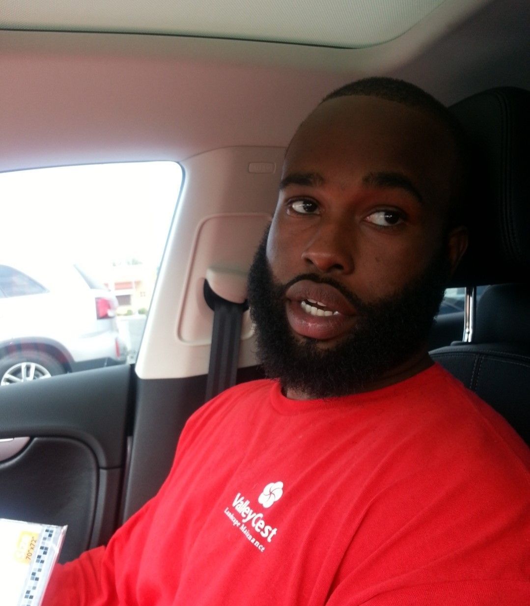 Man seated in a car, wearing a red T-shirt, looking toward the camera.