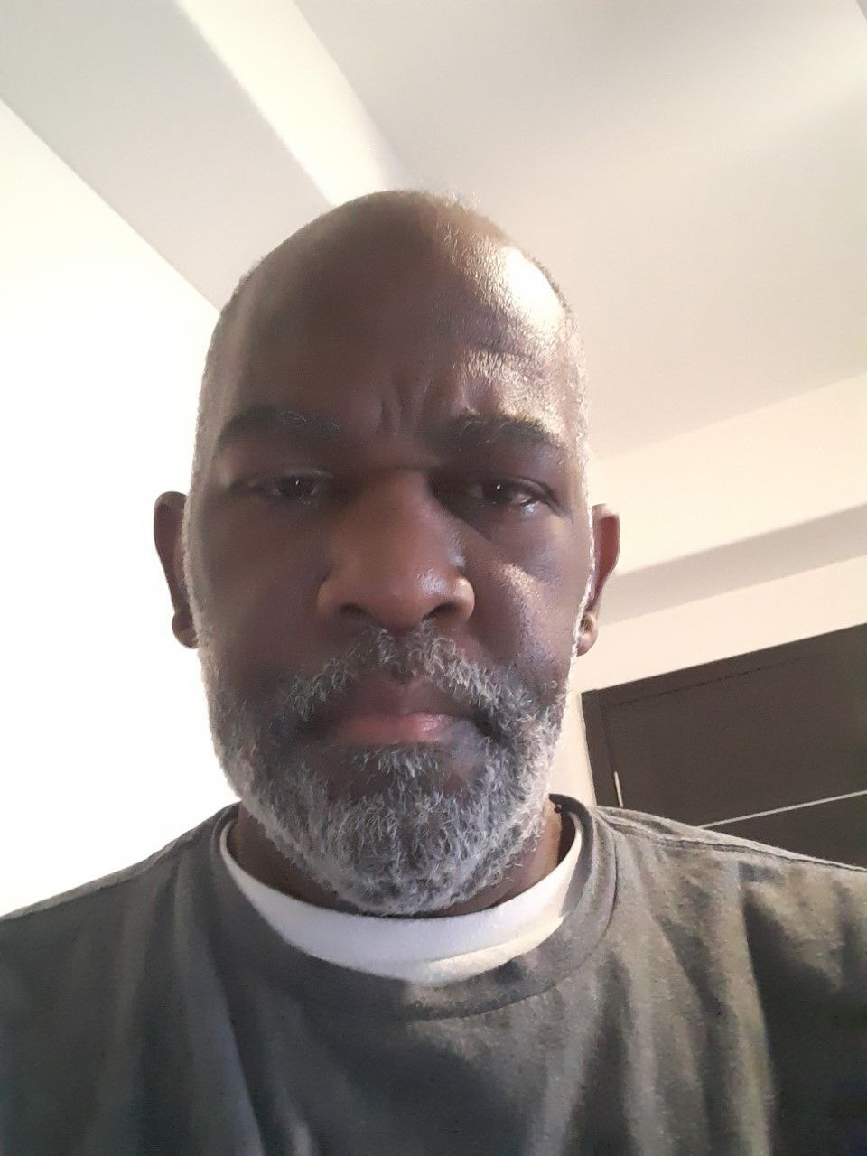 Close-up selfie of a man with a gray beard wearing a dark shirt indoors under soft light