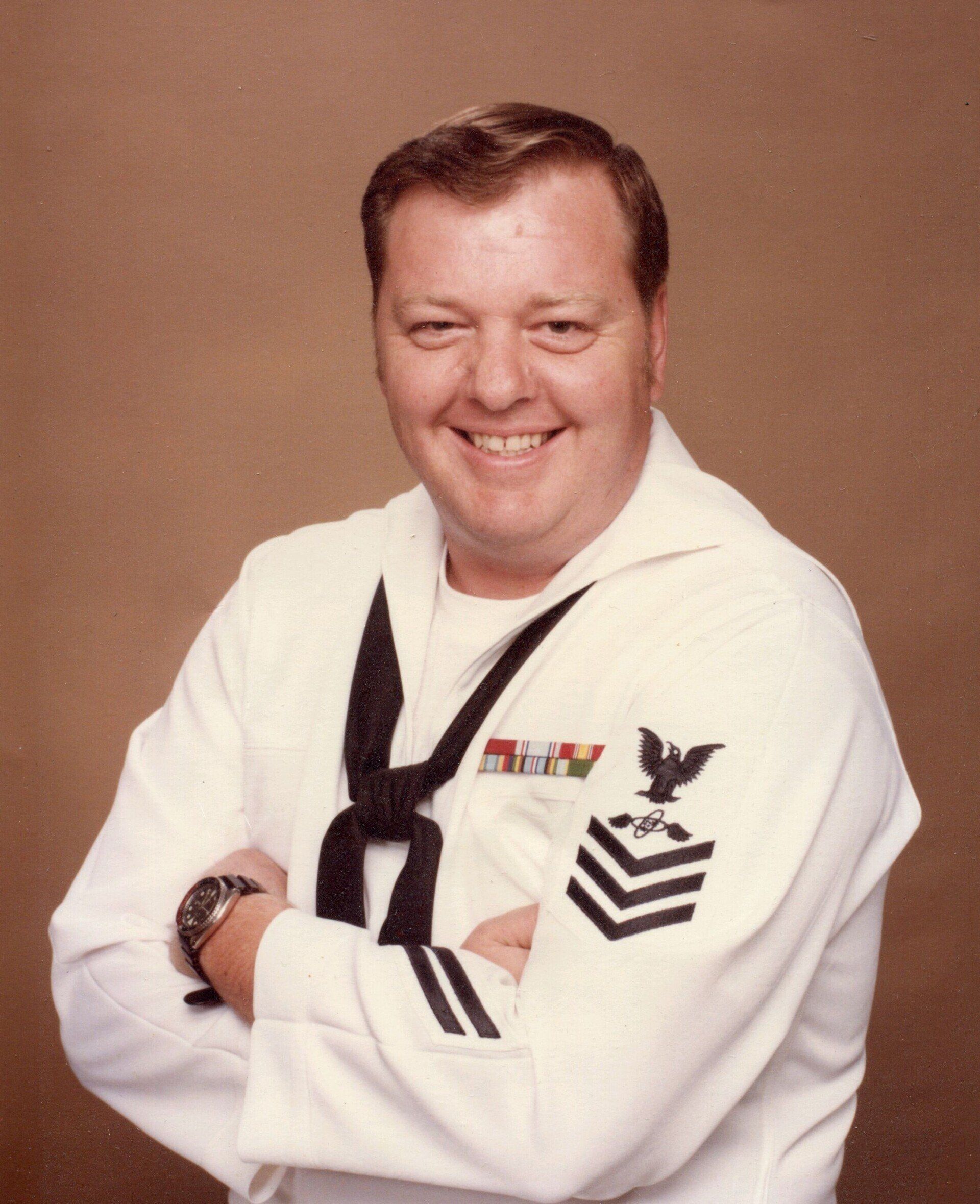 Smiling sailor in white uniform with black neckerchief and insignia, arms crossed against a plain brown background