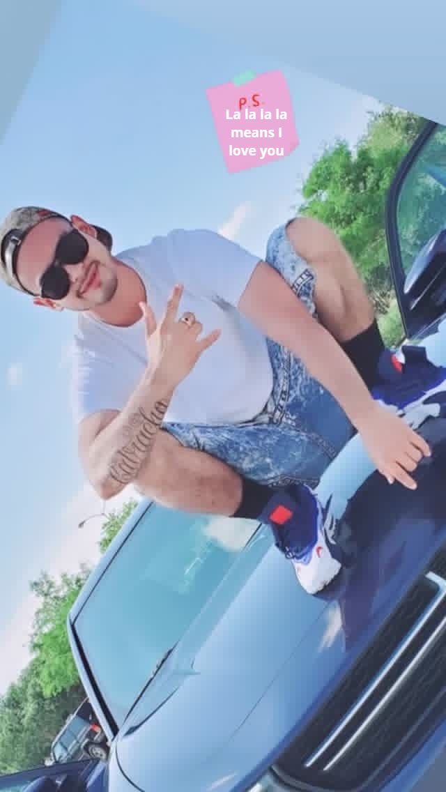 Person posing on the hood of a blue car outdoors under a sunny sky.