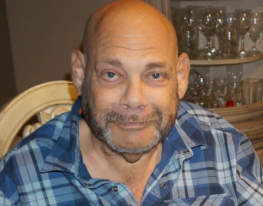 Man in a blue plaid shirt sitting indoors, smiling at the camera.