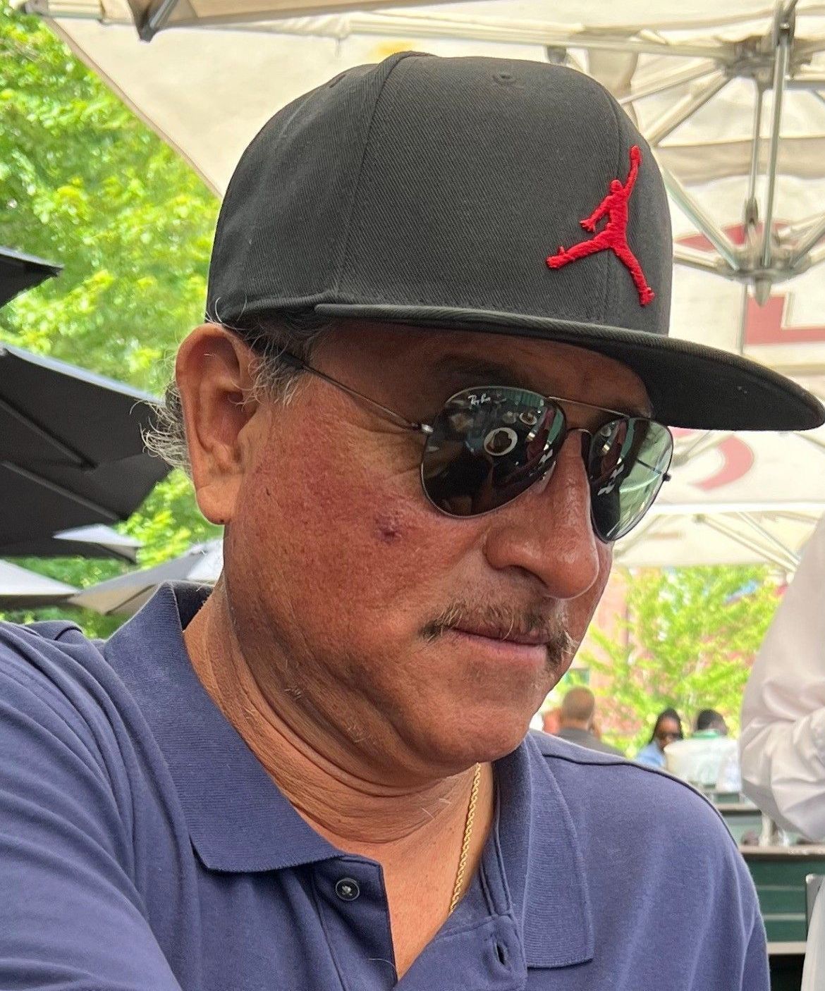 Man in a black cap and sunglasses wearing a navy polo shirt, seated outdoors under a canopy.