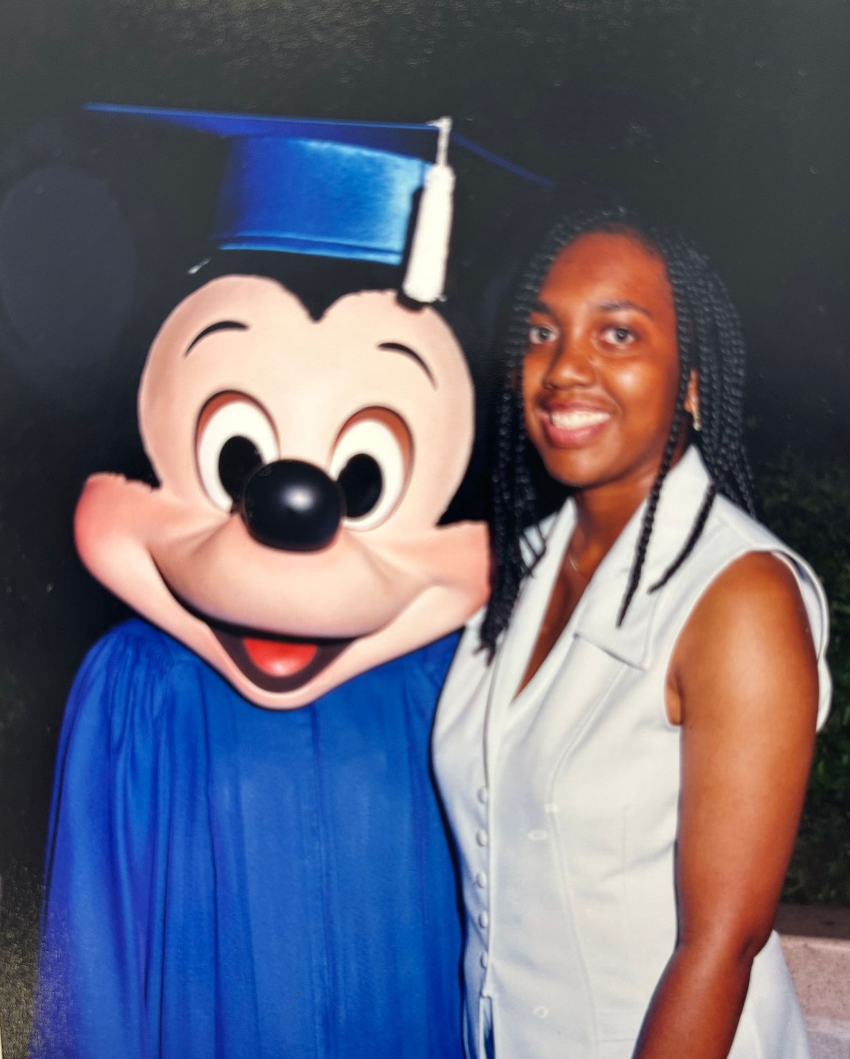 Mickey Mouse in blue graduation cap and gown posing with a smiling woman in a white vest