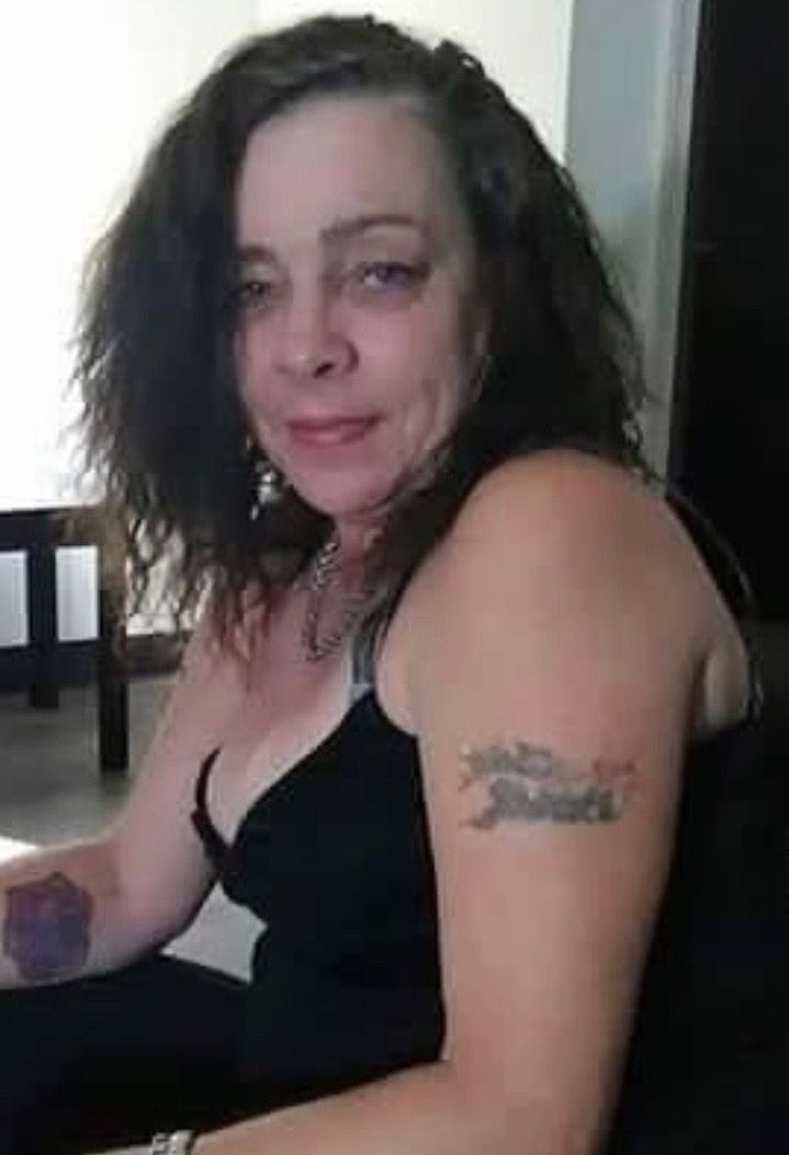 Person seated indoors in a black dress, with wavy dark hair and visible tattoos, looking toward the camera.