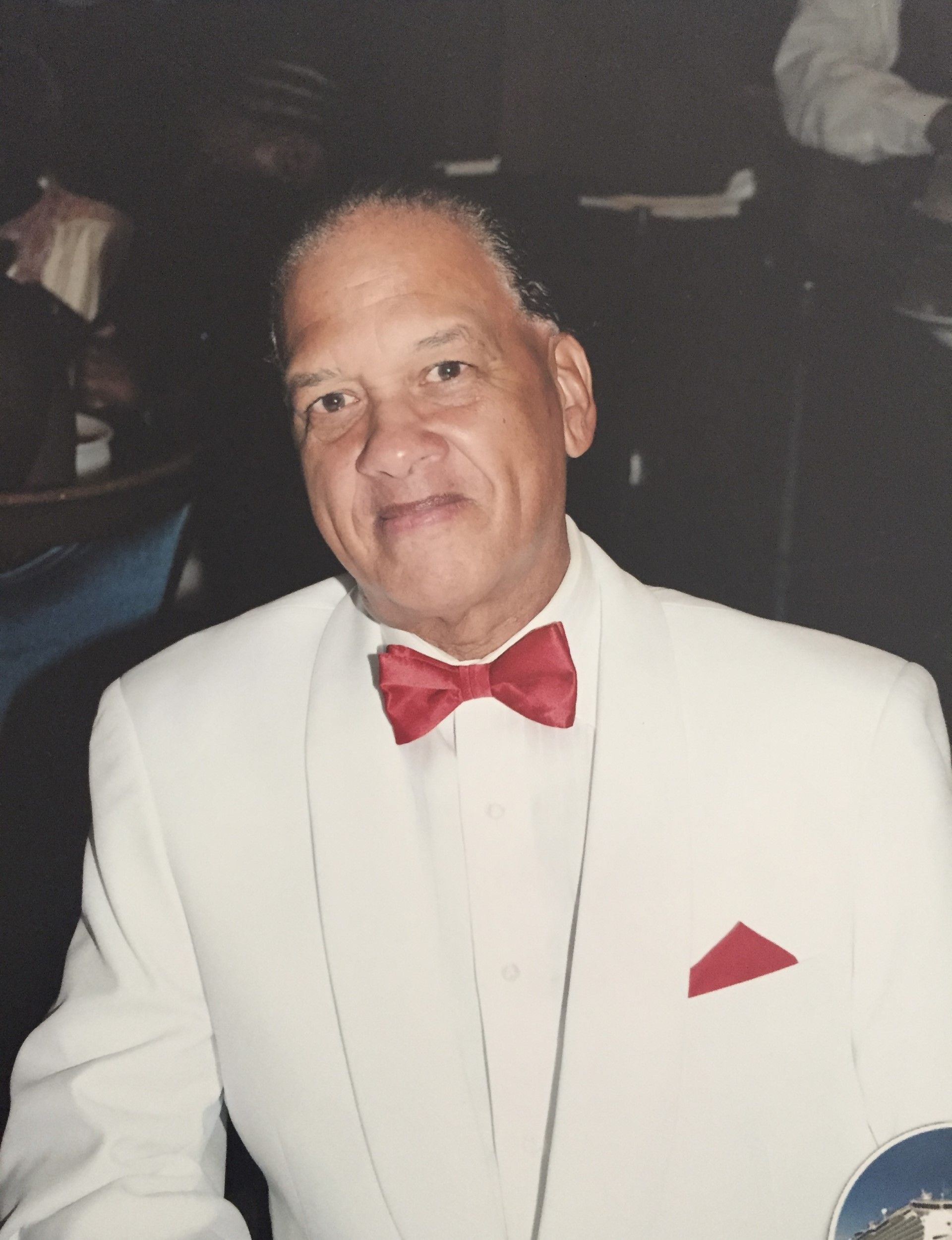 Man in a white tuxedo with red bow tie and pocket square, posing indoors and smiling gently