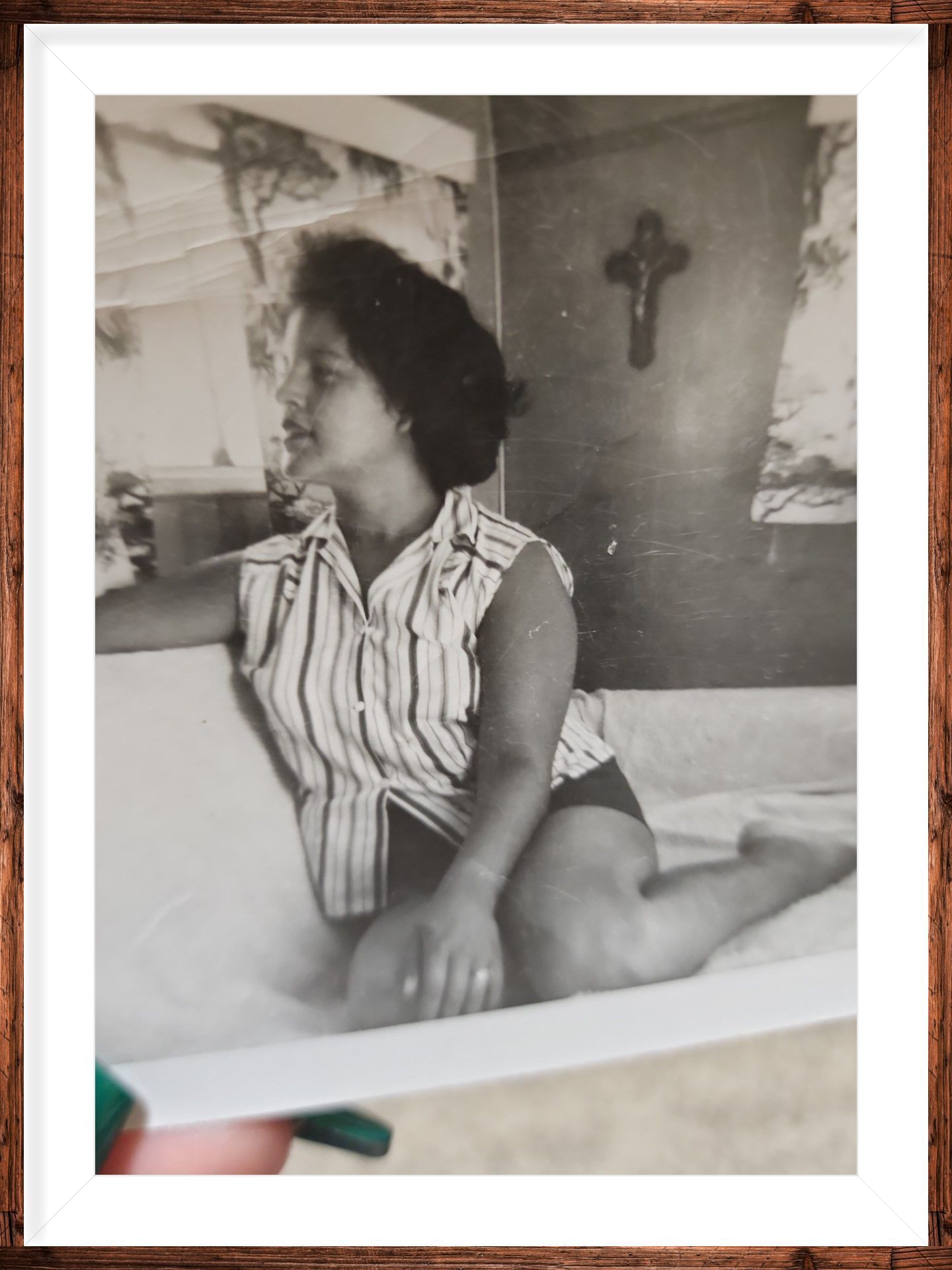 Sepia photo of a person sitting on a bed, looking left, with a cross on the wall behind.