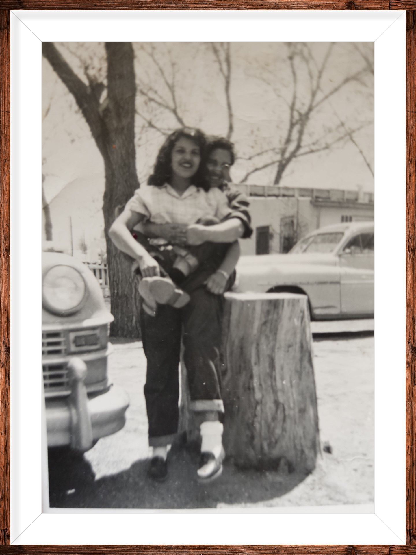 Two children posing outdoors beside a tree stump with parked cars in the background.