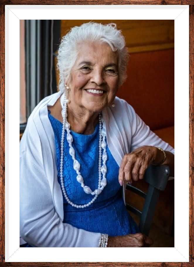 Smiling woman in a blue dress and white cardigan, wearing layered necklaces, seated indoors by a chair