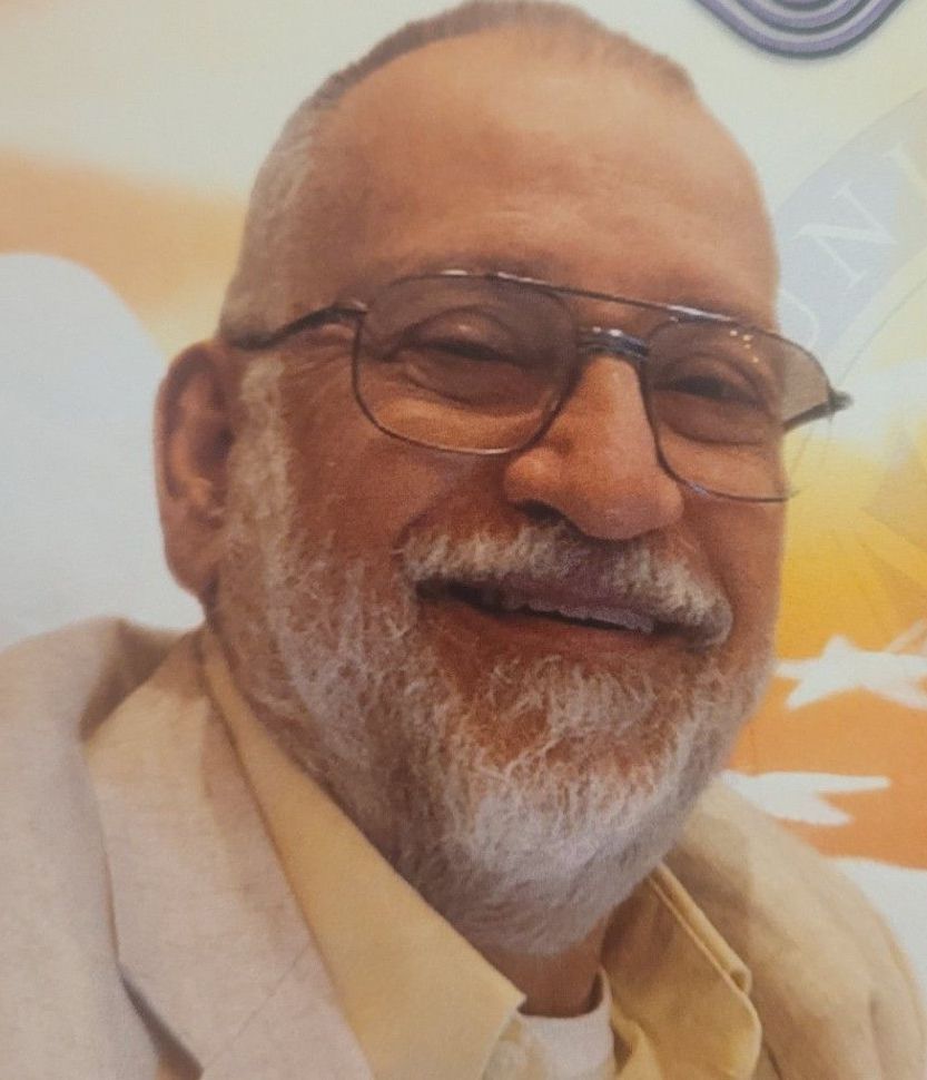 Smiling man with glasses and a white beard, wearing a light beige suit jacket.