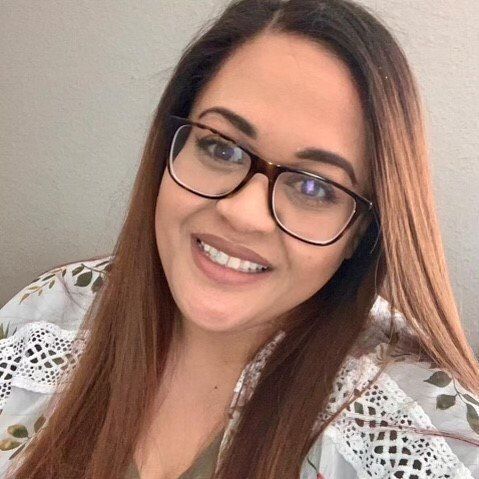 Smiling woman with glasses and long brown hair, wearing a white lace top against a light background