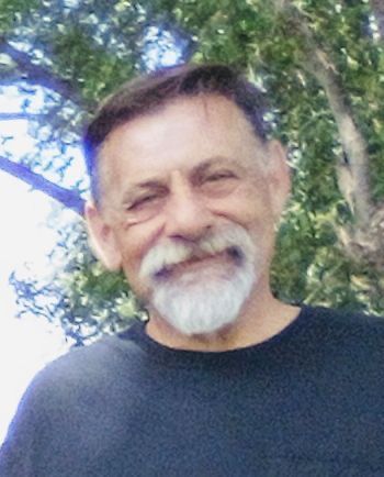 Smiling man with gray beard in a black shirt standing outdoors by trees