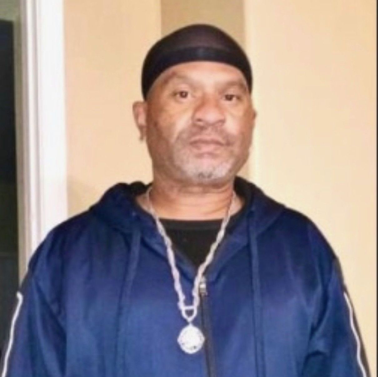 Man standing indoors in a navy hoodie, black head covering, and silver necklace, looking at the camera