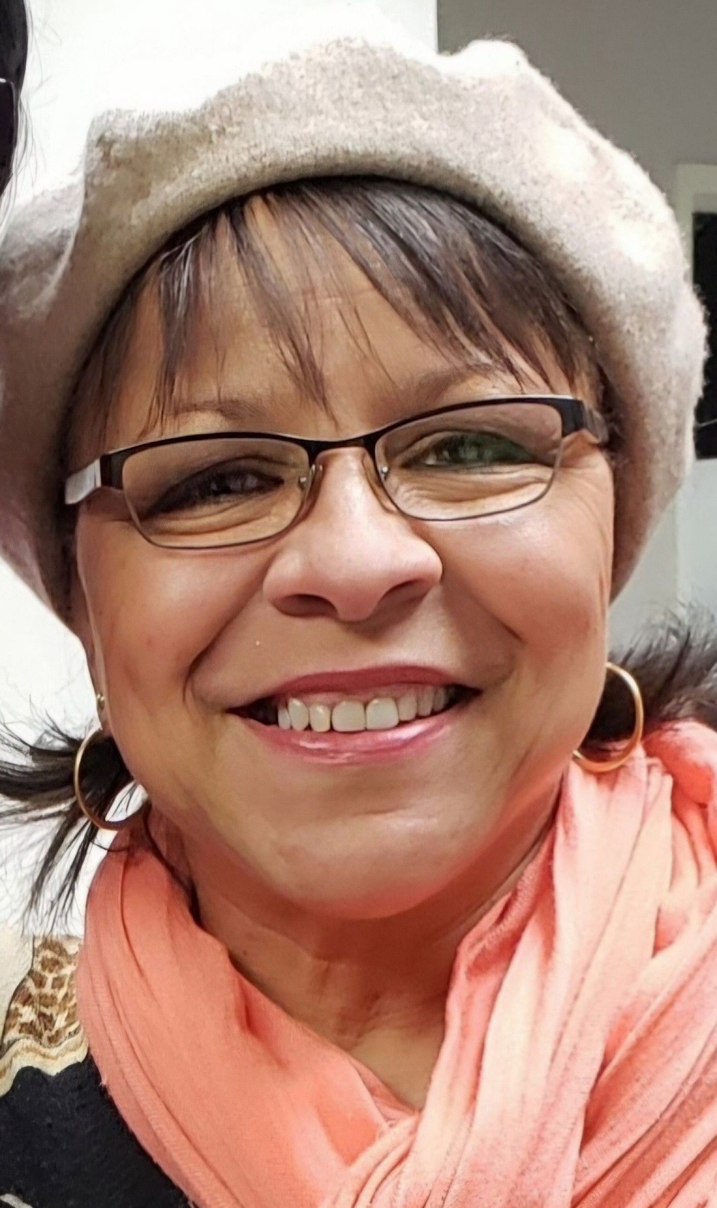 Smiling person wearing glasses, a beige beret, and a coral scarf against a dark background