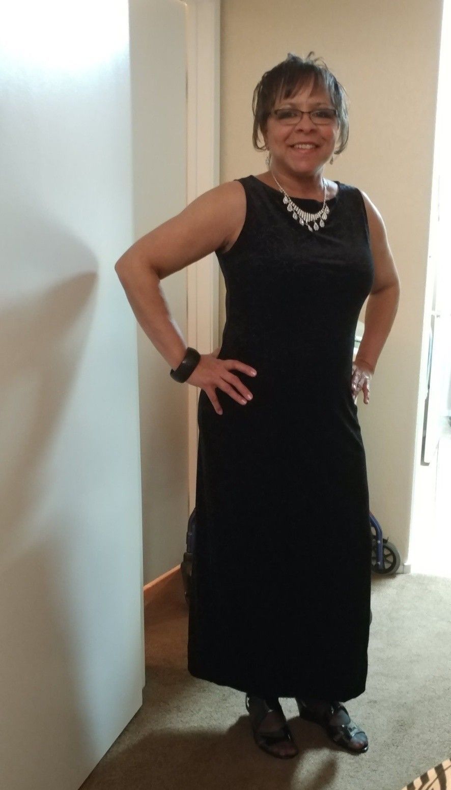 Person in a black dress and necklace standing indoors with hands on hips and smiling.
