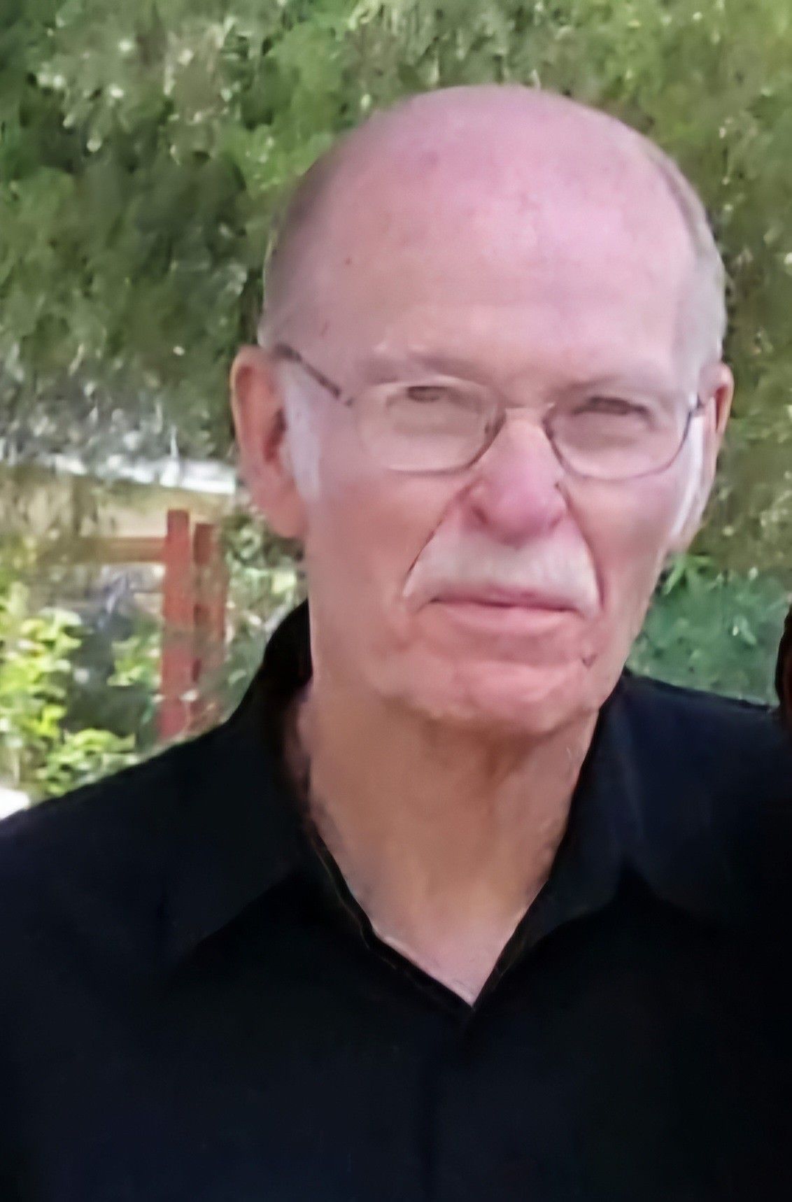 Portrait of a bald man in glasses wearing a black shirt, standing outdoors with trees behind him
