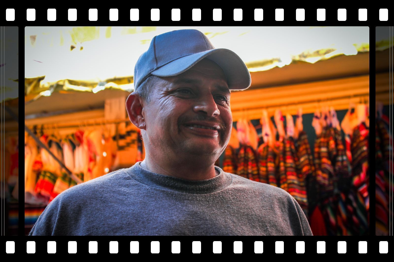 Smiling man in a blue cap and gray sweater standing in a colorful market stall
