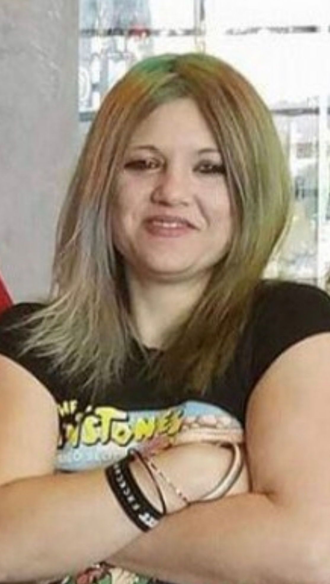 Person with long light-brown hair smiling with arms crossed, wearing a black graphic T-shirt and bracelets.
