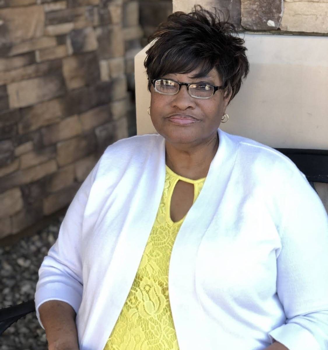 Woman seated indoors wearing glasses, a white cardigan, and a yellow top, against a stone wall.