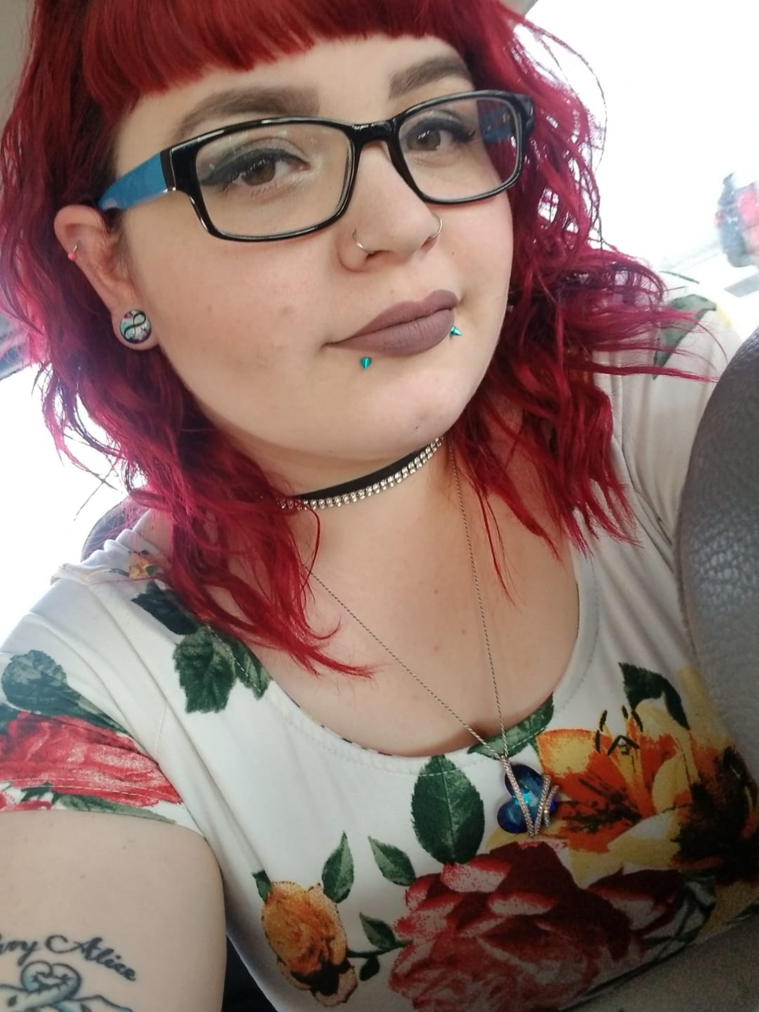 Person with red hair and glasses, wearing a floral top and choker necklace, posing against a bright background.
