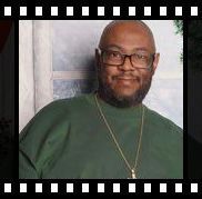Smiling bald man with glasses and a beard, wearing a green shirt and gold necklace