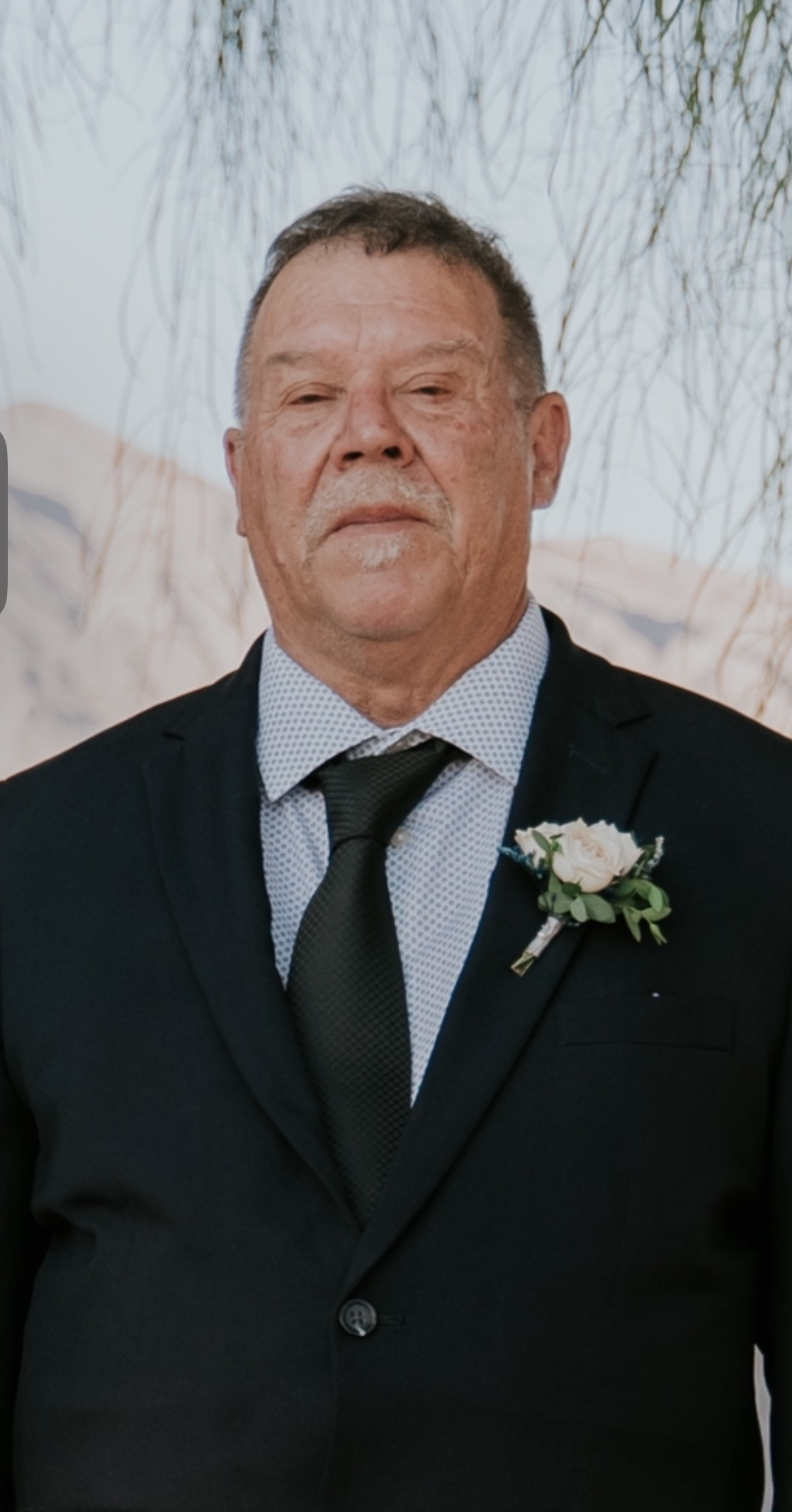 Man in a black suit with a white boutonniere, standing outdoors in front of hanging branches.