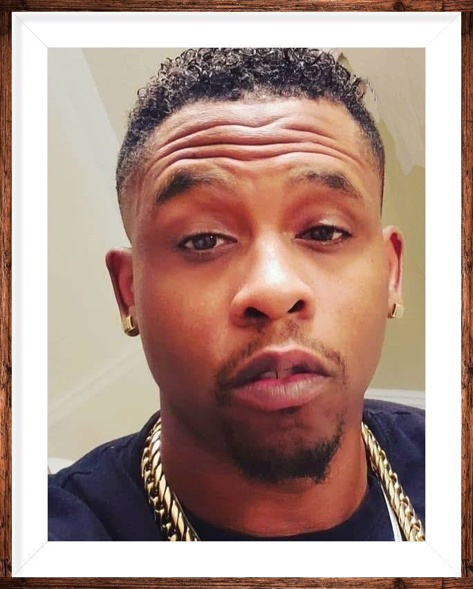 Close-up selfie of a man with short curly hair, gold chains, and a neutral expression against a plain wall