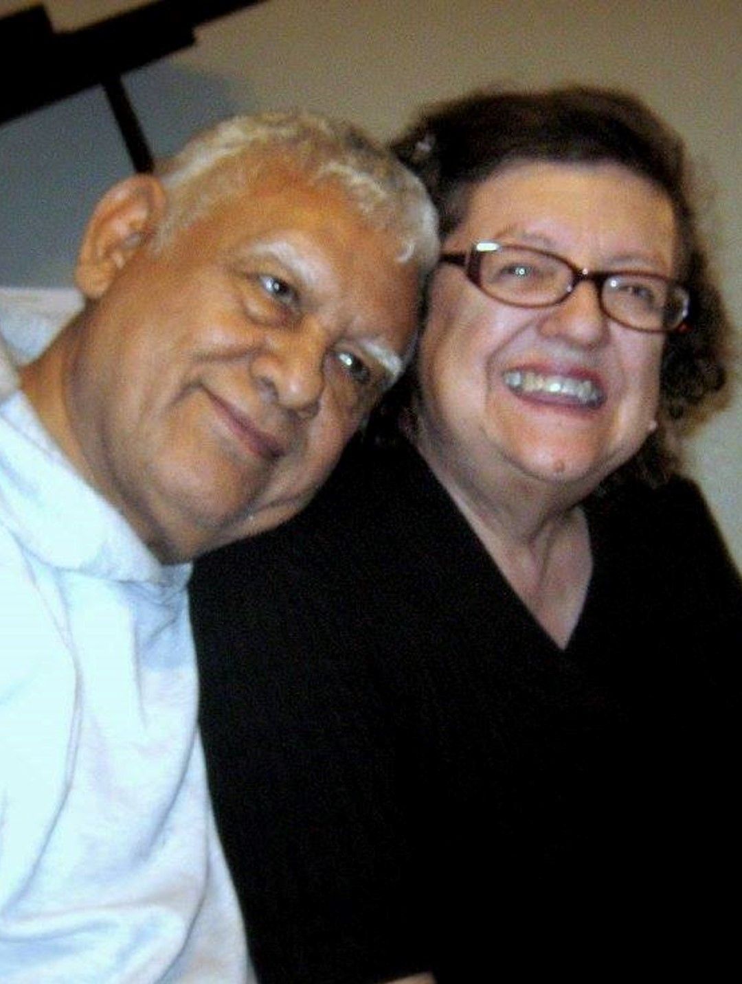 Two people smiling closely together, one wearing glasses and a black top.