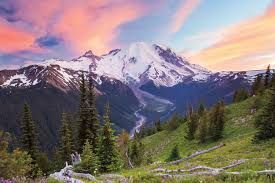 Snowy mountain at sunset above a green alpine valley with pine trees and pink clouds.