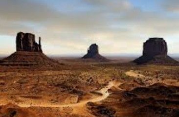 Desert landscape with three towering red rock mesas under a pale blue sky, with a winding dirt path in front