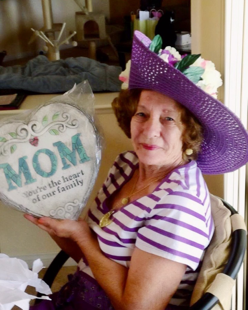 Woman in purple-striped shirt and hat holding a heart-shaped “MOM” gift in a decorated room