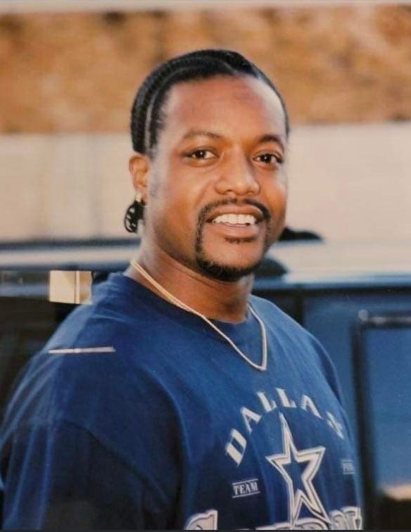 Man in a navy Dallas Cowboys shirt smiling outdoors, with a blurred vehicle behind him