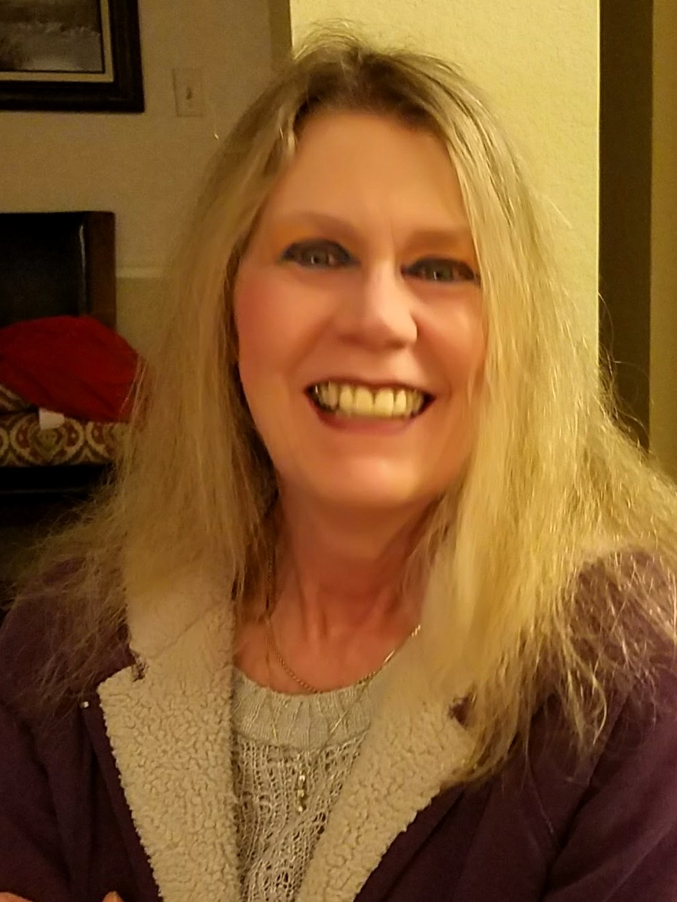 Smiling woman with long blonde hair wearing a purple cardigan and gray top indoors