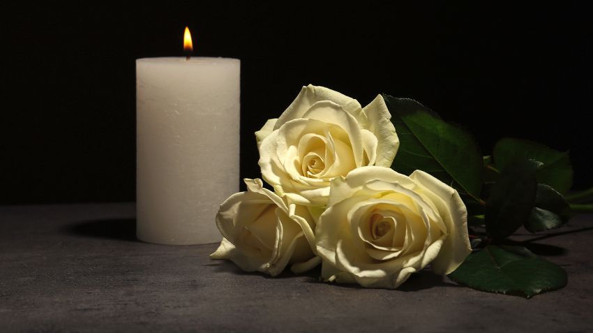 Lit white candle beside three white roses on a dark surface, with green leaves against a black background
